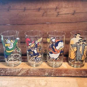 以下が含まれることがあります： カートゥーンキャラクターのイラストが描かれた透明なガラス製の飲み物グラス4つ。グラスには、ウッディー・ウッドペッカー、チリー・ウィリー、バズ・バザード、ハックルベリー・ハウンドが描かれている。