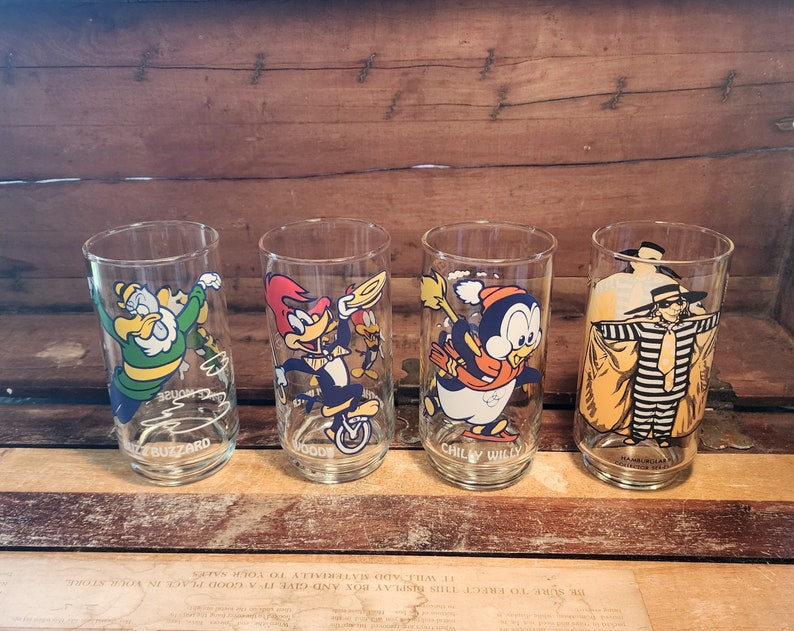 以下が含まれることがあります： カートゥーンキャラクターのイラストが描かれた透明なガラス製の飲み物グラス4つ。グラスには、ウッディー・ウッドペッカー、チリー・ウィリー、バズ・バザード、ハックルベリー・ハウンドが描かれている。