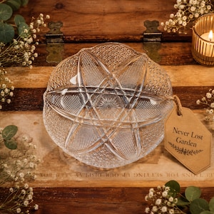 May include: A clear, patterned glass container with a lid sits on a wooden surface. A small tag with the text "Never Lost Garden" hangs from the container. A lit candle in a gold holder and sprigs of white flowers and greenery surround the container.
