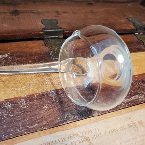 May include: A clear glass gravy boat with a long handle. The boat is sitting on a wooden surface.