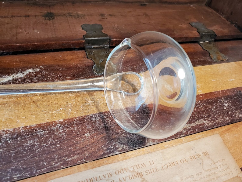 May include: A clear glass gravy boat with a long handle. The boat is sitting on a wooden surface.