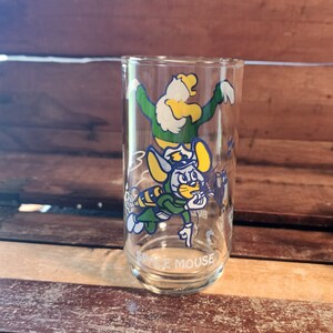 以下が含まれることがあります： 緑色の制服を着た青い帽子をかぶった漫画のネズミと、緑色の制服を着た黄色の鳥の漫画の画像が描かれた透明なガラス製の飲み物グラス。グラスの底には「Space Mouse」と印刷されています。