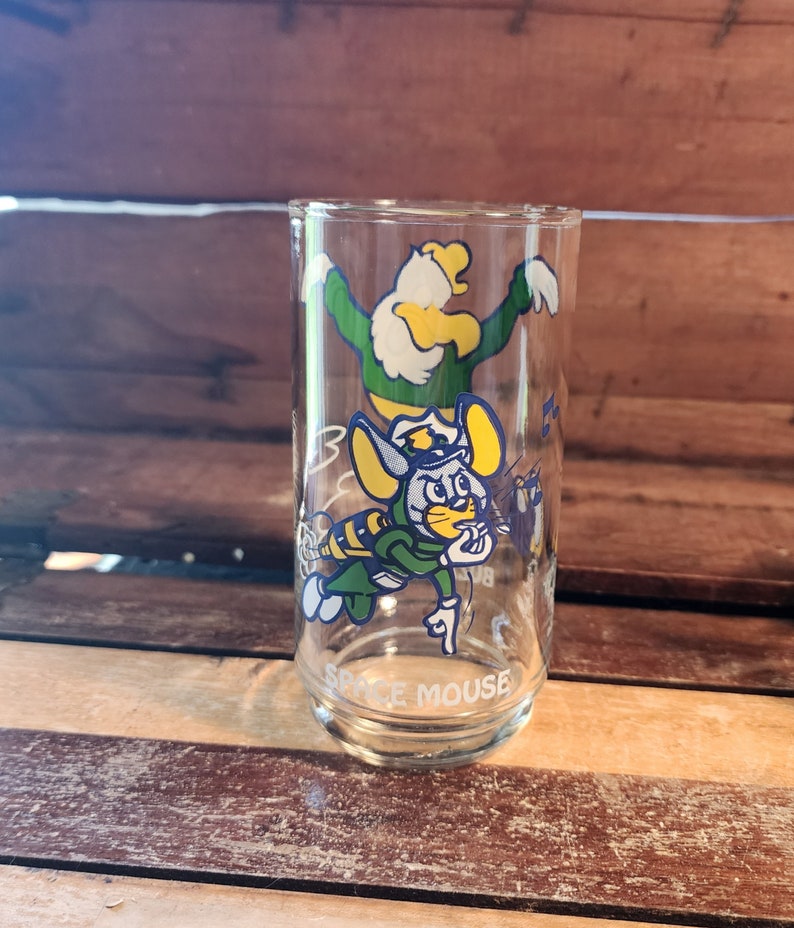 以下が含まれることがあります： 緑色の制服を着た青い帽子をかぶった漫画のネズミと、緑色の制服を着た黄色の鳥の漫画の画像が描かれた透明なガラス製の飲み物グラス。グラスの底には「Space Mouse」と印刷されています。