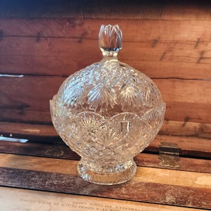 May include: A clear glass candy dish with a domed lid and a cut glass design. The dish has a round base and a decorative knob on the lid.