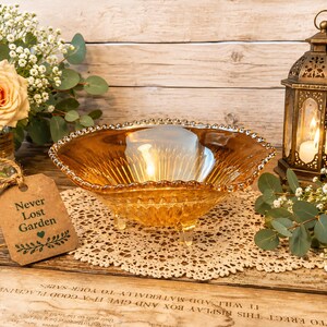 May include: An amber-colored glass bowl with a scalloped rim and three small feet. A lit candle sits inside the bowl. A decorative lantern and a bouquet of flowers are also present. The tag reads "Never Lost Garden".
