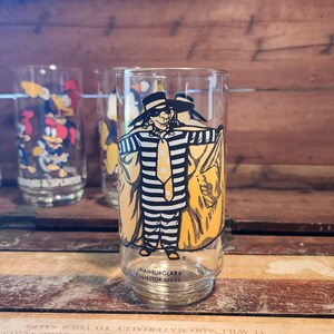 以下が含まれることがあります： マクドナルドのハンバーグラーの黄色と黒のイラストが描かれた透明なガラス。ガラスの底には「Hamburglar&reg; Collector Series」の文字が書かれている。