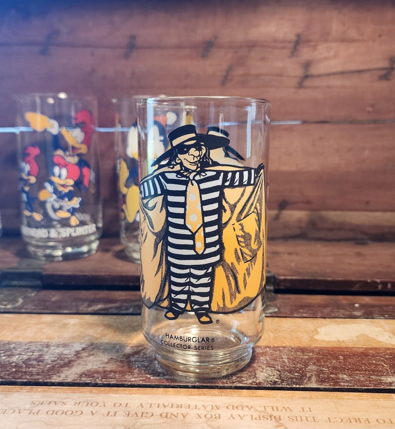以下が含まれることがあります： マクドナルドのハンバーグラーの黄色と黒のイラストが描かれた透明なガラス。ガラスの底には「Hamburglar&reg; Collector Series」の文字が書かれている。