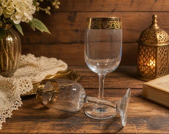 Könnte beinhalten: Zwei klare Weingläser mit goldenen Rändern, eines aufrecht und eines liegend, auf einer Holzoberfläche. Die Gläser haben ein klassisches Design. Eine dekorative goldene Laterne und eine Vase mit Blumen befinden sich im Hintergrund.
