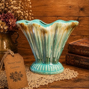 May include: A turquoise ceramic vase with a fluted design and a scalloped rim. The vase has a drip glaze effect with brown and white accents. A tag that says "Never Lost Garden" is attached to a doily.