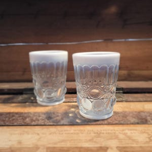 May include: Two vintage clear glass tumblers with a white scalloped rim and decorative etched design. The glasses have a detailed pattern of circles and swirls, perfect for serving beverages. The glasses are displayed on a wooden surface.