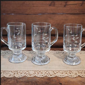 May include: Three clear glass mugs with handles and decorative etched floral designs. The mugs have a pedestal base and are arranged on a beige lace-trimmed cloth. The background is a dark wooden surface.