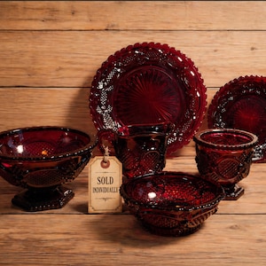 May include: A collection of ruby red glass tableware, including plates, bowls, and glasses, arranged on a wooden surface. The pieces have intricate patterns and textures. A tag reads "SOLD INDIVIDUALLY".