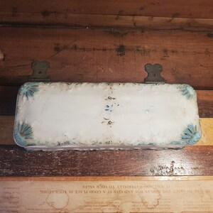 May include: A white porcelain box with blue and white floral designs. The box has a scalloped edge and is decorated with a raised floral pattern. The box is attached to a wooden surface with two metal hinges.