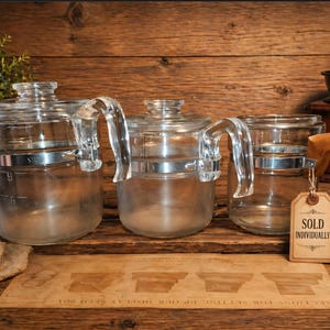 May include: Clear glass coffee pots with handles and metal bands are displayed on a wooden surface. A wooden coffee grinder, a burlap sack of coffee beans, and a wooden scoop with coffee beans are also present. A tag reads "SOLD INDIVIDUALLY."