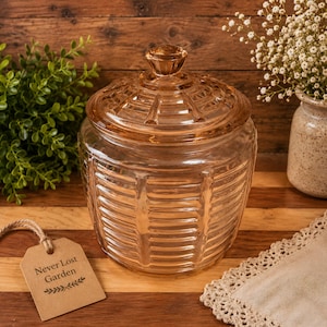 May include: A peach-colored glass jar with a matching lid, featuring a textured horizontal ribbed pattern and a knob handle. A tan rectangular paper tag is attached by a string, printed with the text Never Lost Garden and a leaf graphic.