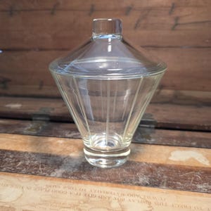 May include: A clear glass jar with a conical shape and a matching lid. The jar has vertical lines etched into the glass, and it sits on a small, round base. The lid has a small, cylindrical handle.
