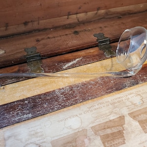 May include: A clear glass ladle with a long, curved handle. The ladle is resting on a wooden surface.