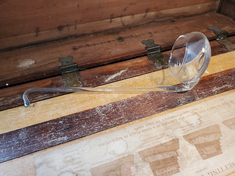 May include: A clear glass ladle with a long, curved handle. The ladle is resting on a wooden surface.