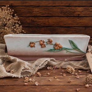 May include: Rectangular ceramic planter with a light pink and white finish, adorned with embossed floral designs in shades of brown and green. The planter is set against a wooden backdrop with dried flowers and a tag that reads "Never Lost Garden."