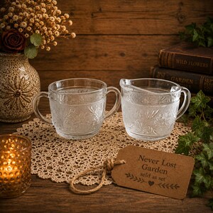 May include: A clear glass sugar bowl with two handles and a matching creamer pitcher, both featuring an embossed floral and stippled pattern. A brown paper tag is attached with twine, printed with the text Never Lost Garden sold as set.