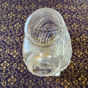 May include: A clear glass container with a wide circular opening at the top and a smaller, rounded extension at the front. The glass features a textured, ribbed pattern throughout its surface, creating a series of vertical lines and ridges.