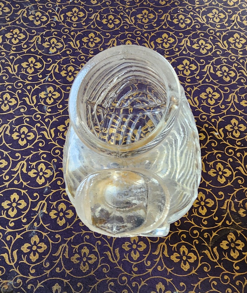 May include: A clear glass container with a wide circular opening at the top and a smaller, rounded extension at the front. The glass features a textured, ribbed pattern throughout its surface, creating a series of vertical lines and ridges.