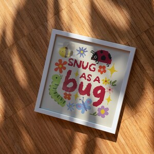 May include: A square white frame containing a cross-stitch embroidery on light fabric. The design features a yellow bee, a red ladybug, a green caterpillar, and a blue butterfly surrounded by colorful flowers and the text SNUG AS A BUG.