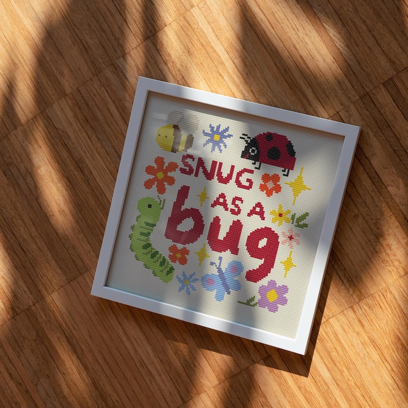 May include: A square white frame containing a cross-stitch embroidery on light fabric. The design features a yellow bee, a red ladybug, a green caterpillar, and a blue butterfly surrounded by colorful flowers and the text SNUG AS A BUG.