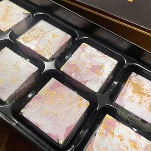 May include: A box of artisan chocolates. Each square chocolate has a white, pink, and gold design. The chocolates are arranged in a black tray with individual compartments. The box is open, revealing the chocolates.