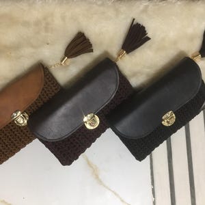 May include: Three crochet clutch purses in brown, dark brown, and black. Each has a flap closure and a gold-coloured clasp. Tassels in matching colours hang from the top. The purses are displayed on a white surface.
