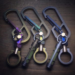 Pode incluir: Três chaveiros de mosquetão de metal em tons de cinza, verde e roxo. Cada chaveiro tem um esquema de cores único e inclui um pequeno acessório cilíndrico com um anel. Os chaveiros são exibidos em uma superfície de madeira.