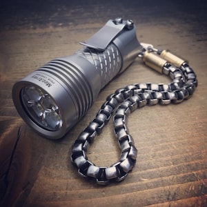 May include: A silver flashlight with a pocket clip and a linked chain wrist strap. The flashlight has a ribbed body and a bright LED light. The wrist strap is made of square metal links. The flashlight is on a wooden surface.