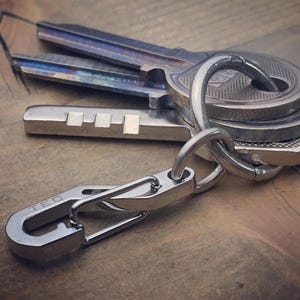 Può includere: Un mazzo di chiavi su un portachiavi argentato con un moschettone. Le chiavi sono di varie forme e dimensioni, con finiture argentate e iridescenti. Il portachiavi e il moschettone sono in metallo lucido.