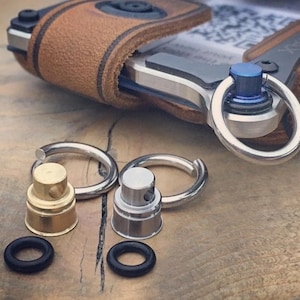 Puede incluir: Primer plano de una funda de teléfono de cuero marrón con un accesorio de anillo plateado y azul. También se ven dos anillos plateados, dos accesorios de botella de spray de metal y dos juntas tóricas de goma negra. Sobre una superficie de madera.