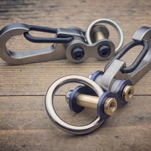 May include: Two metal carabiner keychains with a silver finish and black accents. The keychains have a ring and a clasp, with a small chain-like connector. The keychains are on a wooden surface.