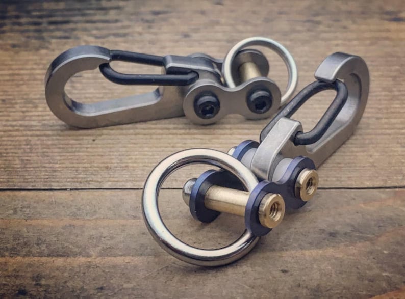 May include: Two metal carabiner keychains with a silver finish and black accents. The keychains have a ring and a clasp, with a small chain-like connector. The keychains are on a wooden surface.