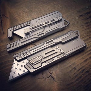 Puede incluir: Dos cuchillos utilitarios plateados con diseños intrincados. Un cuchillo presenta una hoja con un patrón de estrellas, mientras que el otro tiene una hoja lisa. Ambos cuchillos tienen un clip y una serie de pequeños agujeros y crestas.