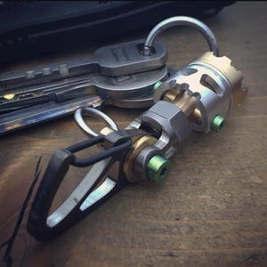 May include: A silver and gold-toned keychain with a mechanical design, featuring a carabiner clip and a ring holding several keys. The keychain has green screws and brass-coloured accents. The background is a wooden surface.