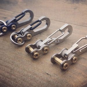 May include: Four metal keychains with carabiner clips and chain link details. The keychains are silver and gold in color, with a blue accent on one. They are arranged on a wooden surface.
