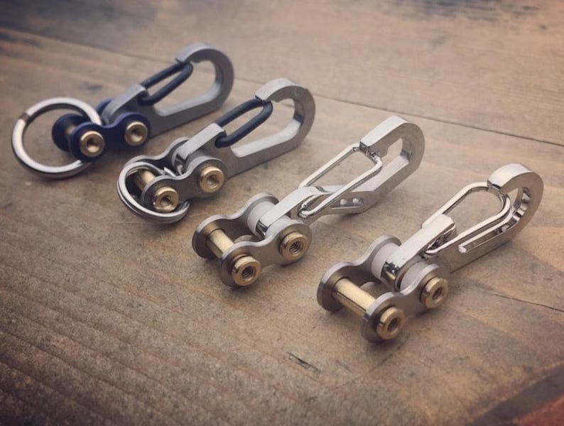 May include: Four metal keychains with carabiner clips and chain link details. The keychains are silver and gold in color, with a blue accent on one. They are arranged on a wooden surface.