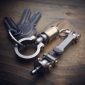 May include: Close-up of a set of keys and two metal keychains. One keychain has a cylindrical brass and silver design, and the other has a silver and brass hinged design. The items are on a wooden surface.