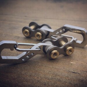 May include: Two silver-colored keychains with a unique design. Each keychain features a carabiner-style clip and a chain-link section with brass-colored accents. The keychains are displayed on a wooden surface.