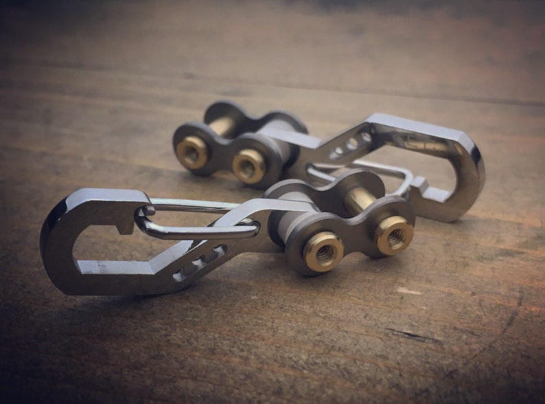 May include: Two silver-colored keychains with a unique design. Each keychain features a carabiner-style clip and a chain-link section with brass-colored accents. The keychains are displayed on a wooden surface.