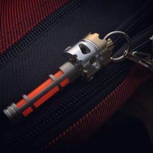 Puede incluir: Un llavero metálico con un diseño cilíndrico. El llavero presenta una parte superior plateada y dorada con un anillo para sujetar llaves. La sección central es plateada con un diseño rectangular naranja. El llavero descansa sobre una superficie texturizada negra y roja.