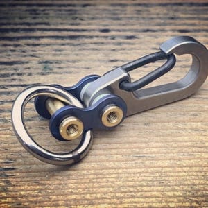 May include: A metal keychain with a carabiner clip and a ring. The keychain has a blue and silver design with gold-colored accents. The item is shown on a wooden surface.