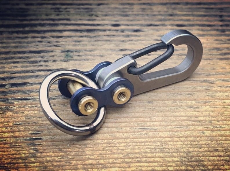 May include: A metal keychain with a carabiner clip and a ring. The keychain has a blue and silver design with gold-colored accents. The item is shown on a wooden surface.