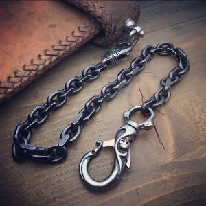 Könnte beinhalten: Eine silberfarbene, robuste Geldbörsenkette mit einem Verschluss im Schädel-Design und dekorativen Gliedern. Die Kette wird auf einer Holzoberfläche neben einer braunen Lederbrieftasche präsentiert.