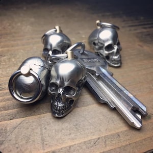 Puede incluir: Una colección de llaveros de calavera plateados y una llave, dispuestos sobre una superficie de madera. Los llaveros presentan diseños detallados de calaveras con anillas. La llave es plateada y tiene forma estándar.