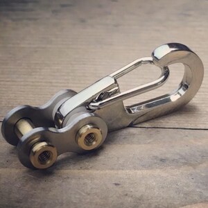May include: A silver-colored keychain with a unique design, featuring a carabiner-style clip and a chain link section. The chain links have gold-colored accents. The keychain is made of metal and rests on a wooden surface.
