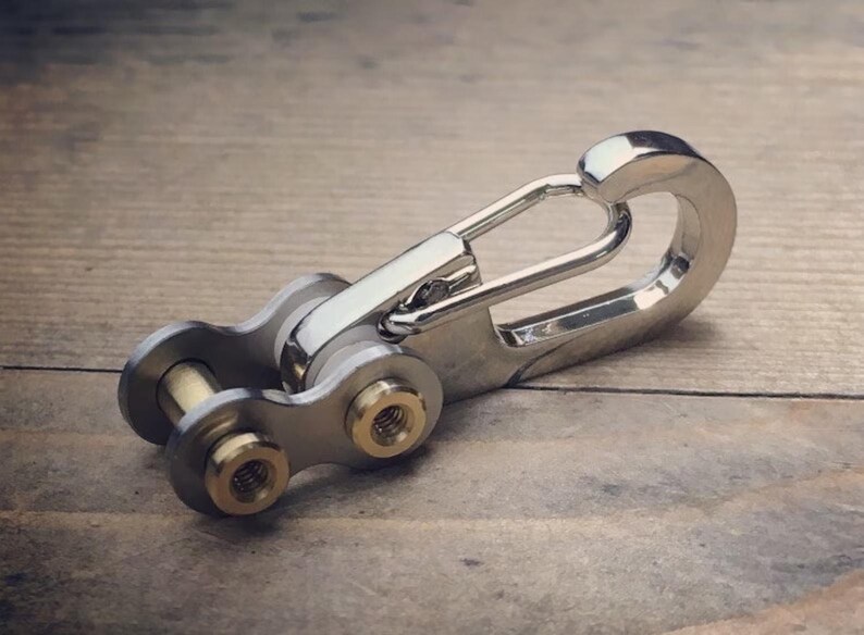 May include: A silver-colored keychain with a unique design, featuring a carabiner-style clip and a chain link section. The chain links have gold-colored accents. The keychain is made of metal and rests on a wooden surface.